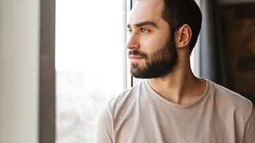 Handsome serious young bearded man standing at the window at home, looking away